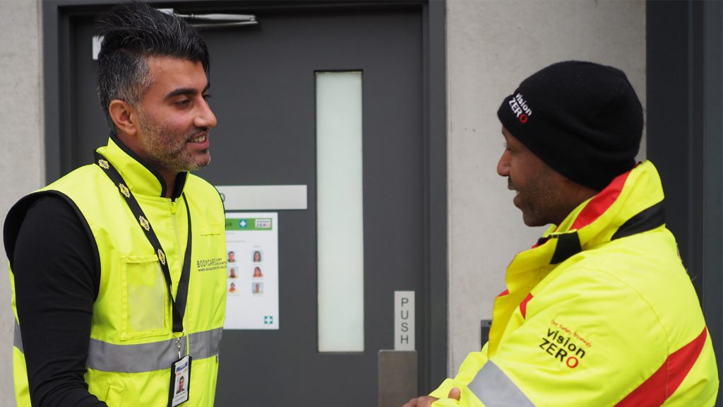 A physiotherapist in a high-vis vest speaking with a worker at a job site.